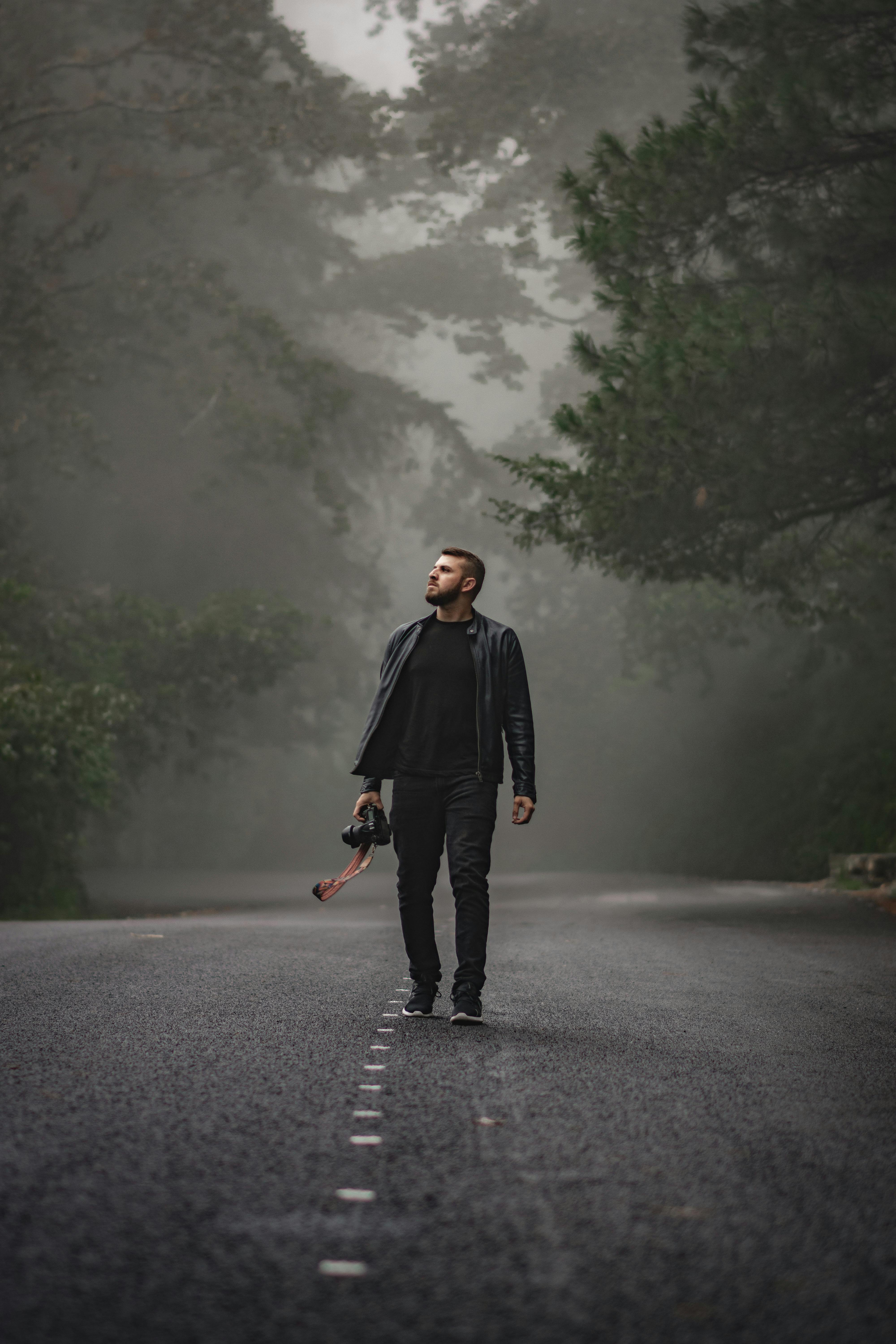 A man dressed in black walks down a misty forest road with a camera in hand, surrounded by towering trees and a mysterious foggy atmosphere.