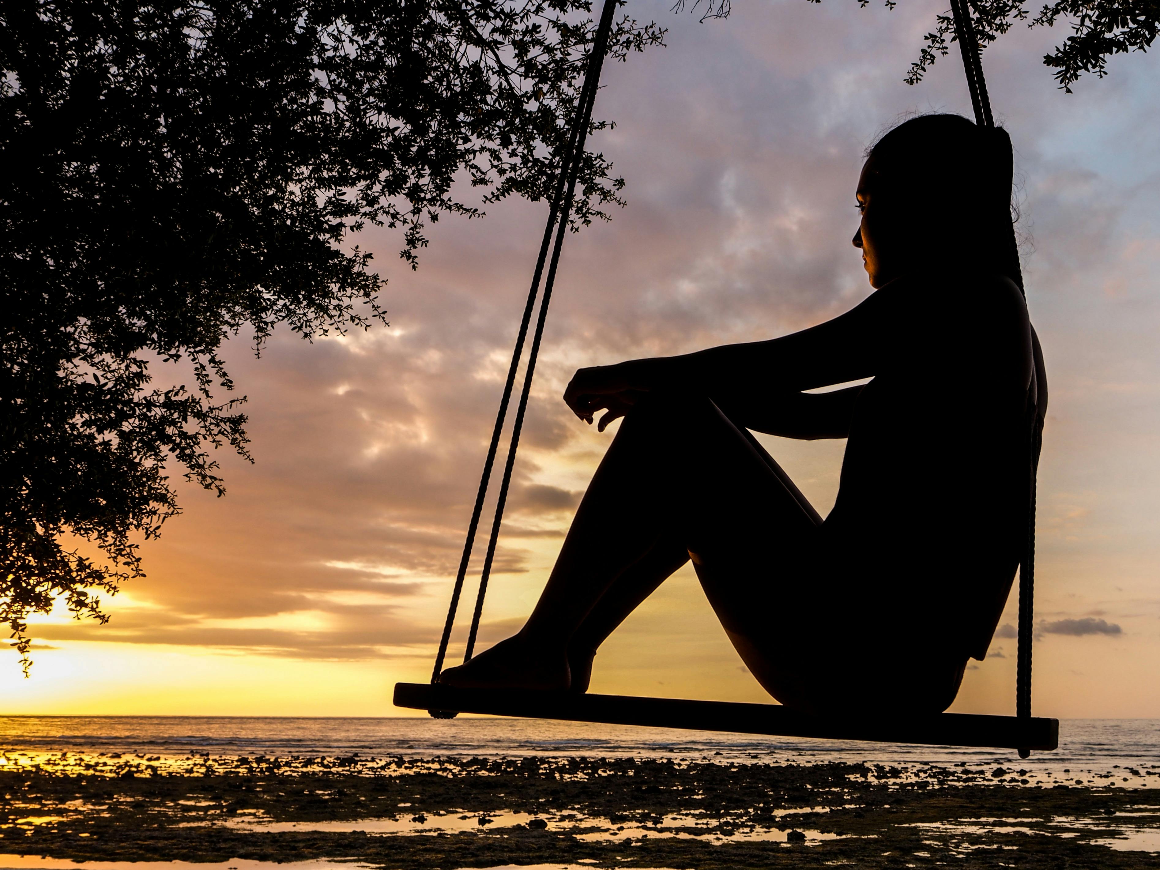 A peaceful silhouette of a woman on a swing, gazing at a golden sunset over a tranquil beach — a serene moment of reflection and calm