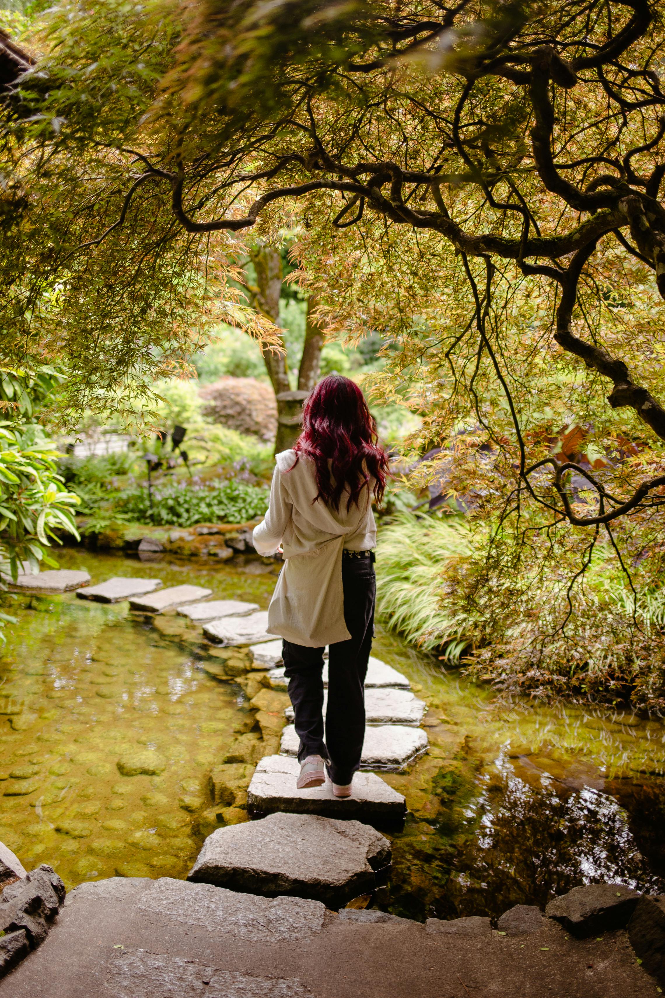 A woman walks across a stone path over a clear stream in a peaceful garden, surrounded by lush foliage and gently arching trees.